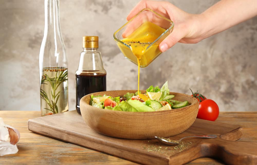 Hand Pouring Mustard Into A Bowl Of Fresh Salad On The Table